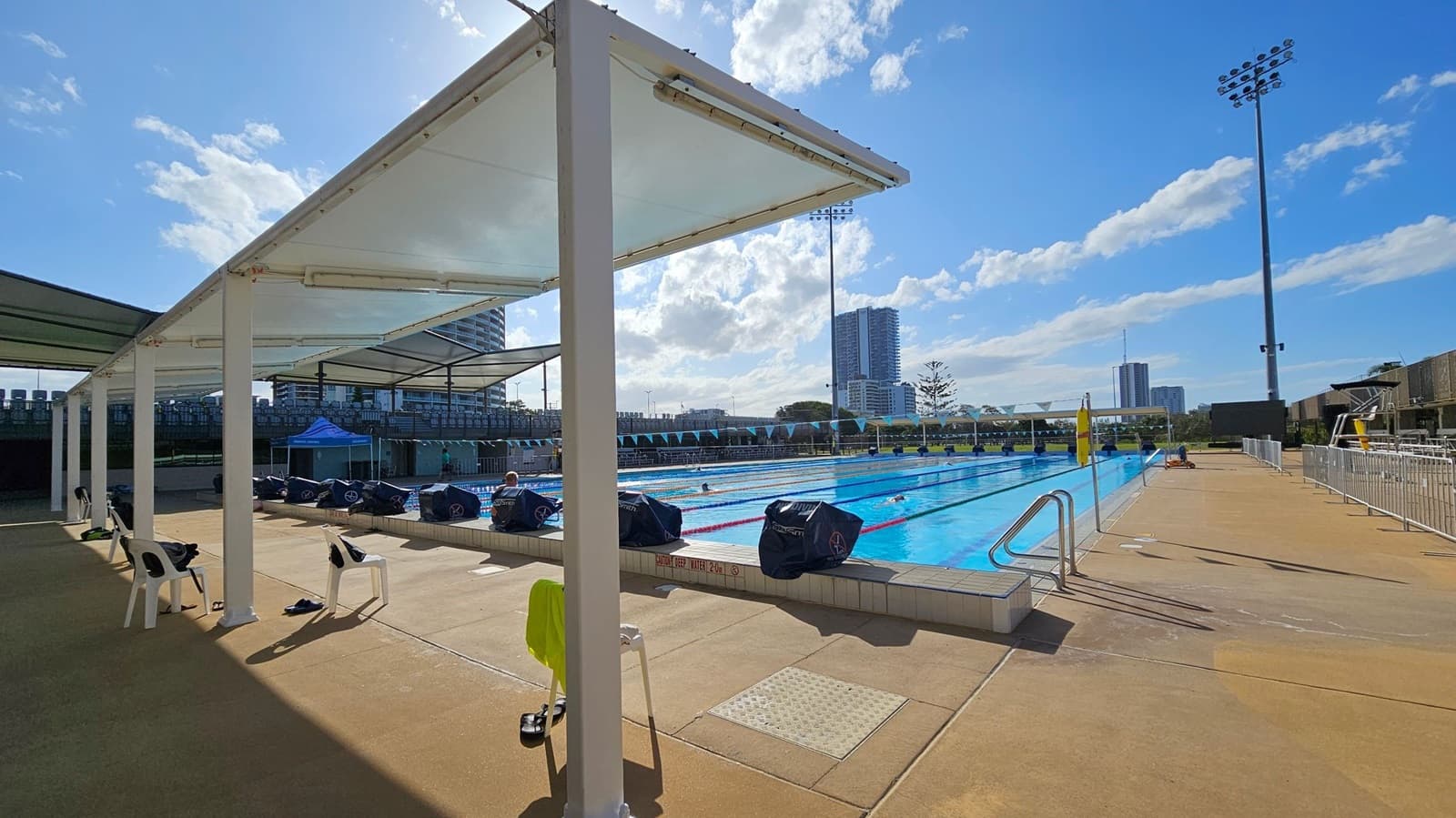 An outdoor swimming pool with lanes is on the right, surrounded by a concrete deck. On the left, there are white chairs and a shaded area with a canopy supported by white pillars. Several bags and personal items are placed on the chairs and ground. In the background, tall buildings and a few trees are visible. Floodlights are positioned around the pool area. The sky is partly cloudy.