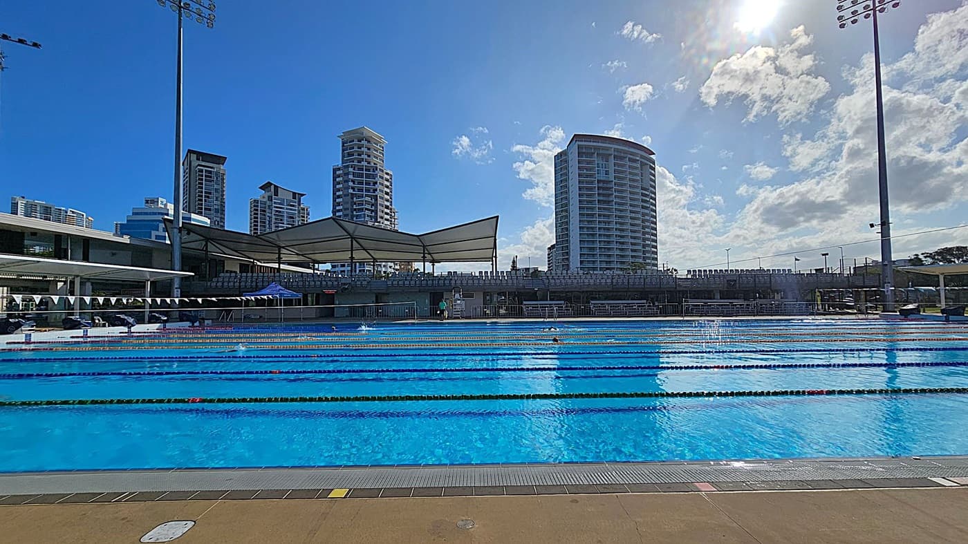 An outdoor swimming pool with multiple lanes is in the foreground, surrounded by a concrete deck. In the background, there are several tall buildings with modern architecture. A large canopy structure provides shade over seating areas. Tall floodlights are positioned on both sides of the pool. The sky is clear with some clouds, and the sun is visible, casting reflections on the water.
