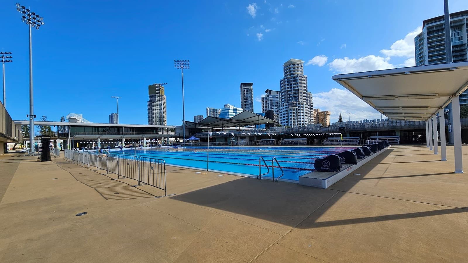 An outdoor swimming pool with clear blue water is in the centre, surrounded by a concrete deck. On the right, there is a row of white canopies providing shade. On the left, tall floodlights and a metal fence line the pool area. In the background, several high-rise buildings are visible against a blue sky. The pool has lane dividers and starting blocks, and the area is well-lit with overhead lights.