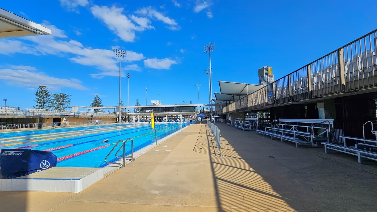 An outdoor swimming pool with multiple lanes is on the left, bordered by a tiled edge. The pool area is surrounded by metal railings and has a lifeguard chair. On the right, there are shaded seating areas with metal benches and a walkway. Tall floodlights and a few trees are visible in the background, along with a tall building. The ground is a mix of concrete and tiled surfaces.