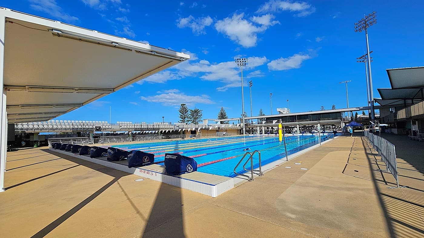 An outdoor swimming pool with multiple lanes, surrounded by a concrete deck. On the left, there's a shaded area with a white canopy. The right side has a metal fence and a pathway. In the background, there are spectator stands with rows of seats and tall floodlights. A few trees are visible beyond the stands. The pool has starting blocks at one end, and a ladder on the right side.
