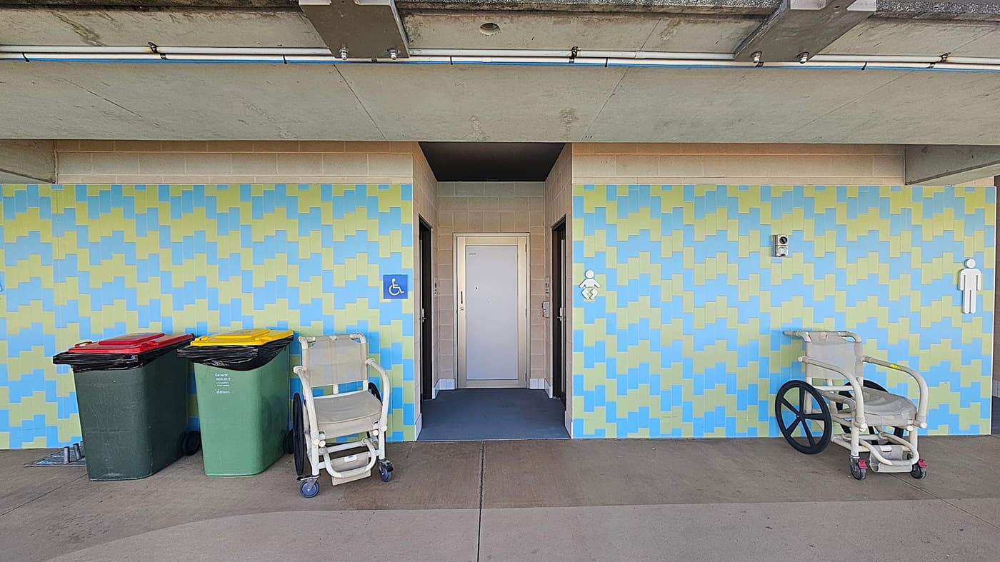 A public restroom entrance with a patterned wall in blue and yellow. In the centre, there's a door with a small ramp leading to it. On the left, two large bins with red and yellow lids are next to a wheelchair. On the right, another wheelchair is positioned near a male restroom sign. The ceiling is concrete with visible pipes and fixtures. The floor is a smooth, grey surface.