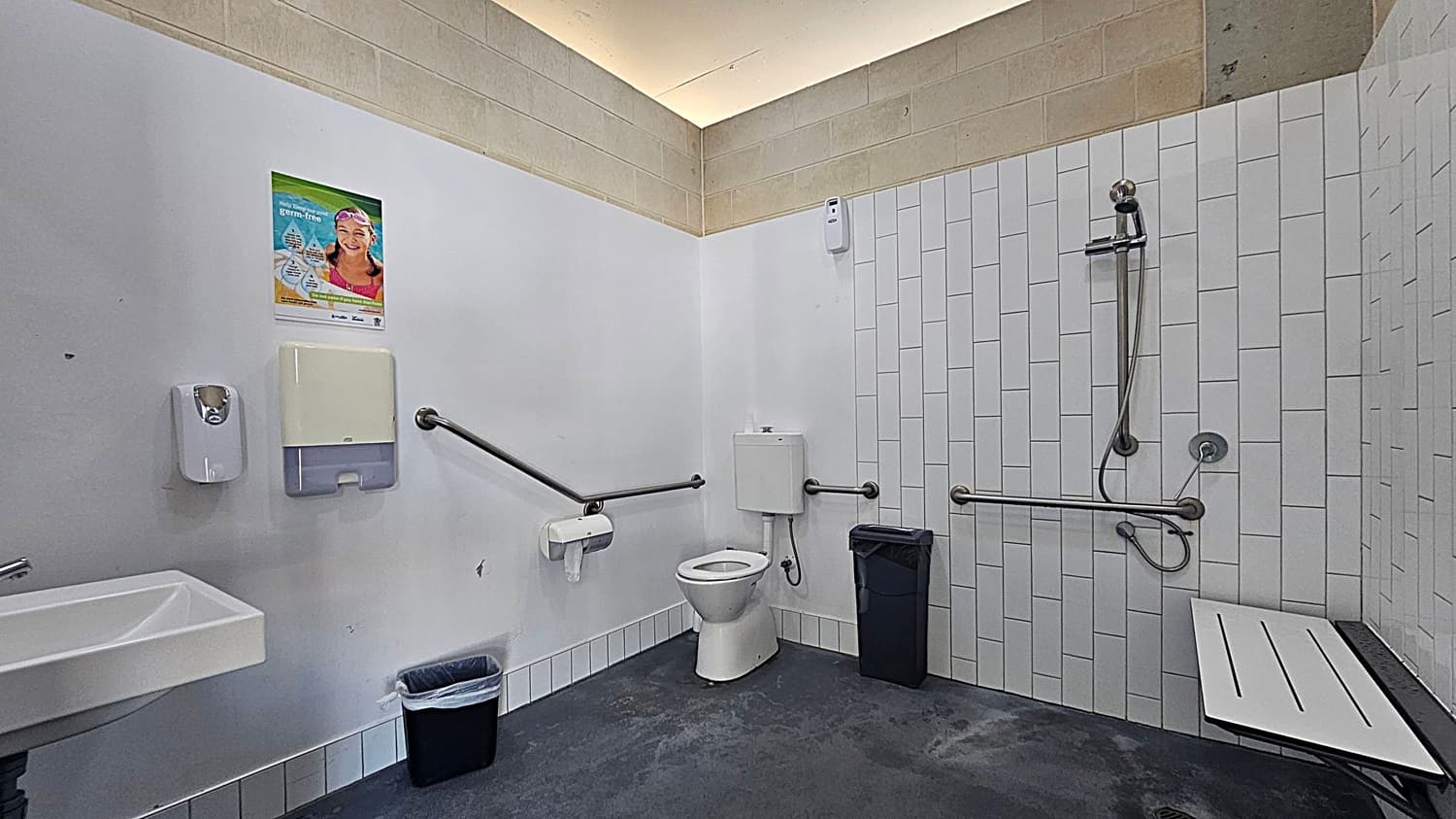 A public accessible bathroom with a white tiled wall on the right featuring a shower with a handheld showerhead and grab bars. A foldable shower seat is attached to the wall. In the centre, there is a toilet with grab bars on both sides and a bin nearby. On the left, a sink with a soap dispenser and paper towel holder is mounted on the wall. A poster is displayed above the paper towel holder. The floor is dark grey, and the ceiling has a light strip along the edge.