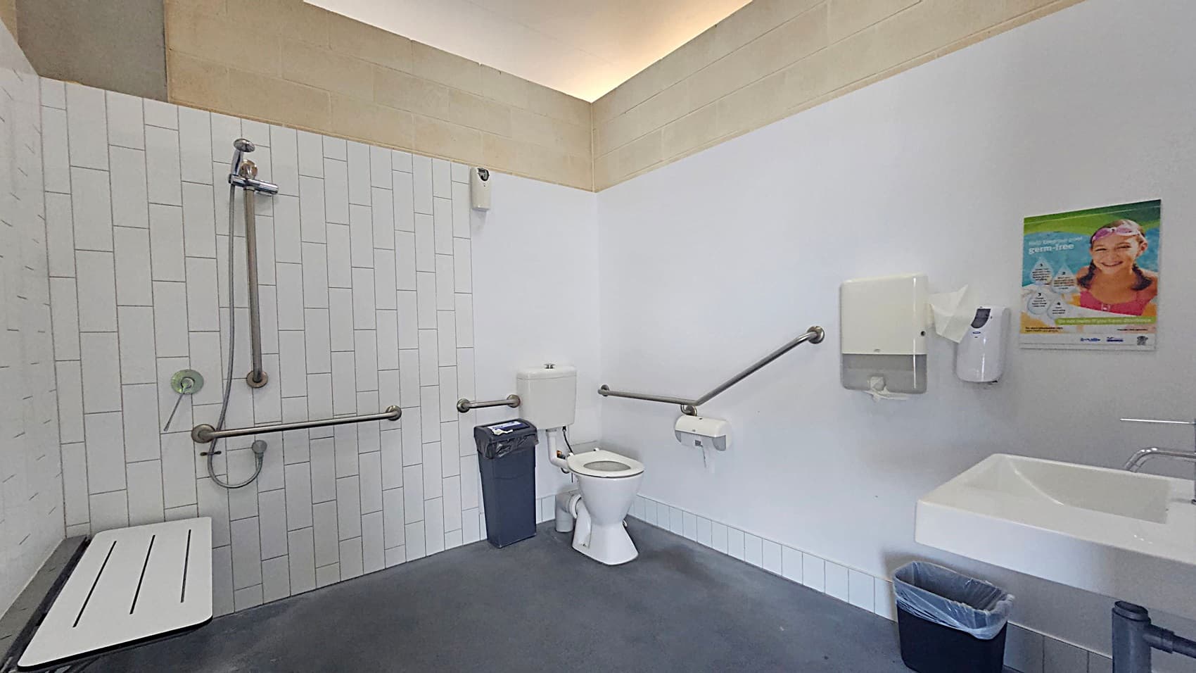A spacious accessible bathroom with a shower area on the left, featuring a wall-mounted showerhead, grab bars, and a foldable seat. In the centre, there's a toilet with grab bars on both sides and a bin nearby. On the right, a sink with a soap dispenser and a hand dryer above it. A poster is mounted on the wall. The floor is grey, and the ceiling has a light-coloured border.