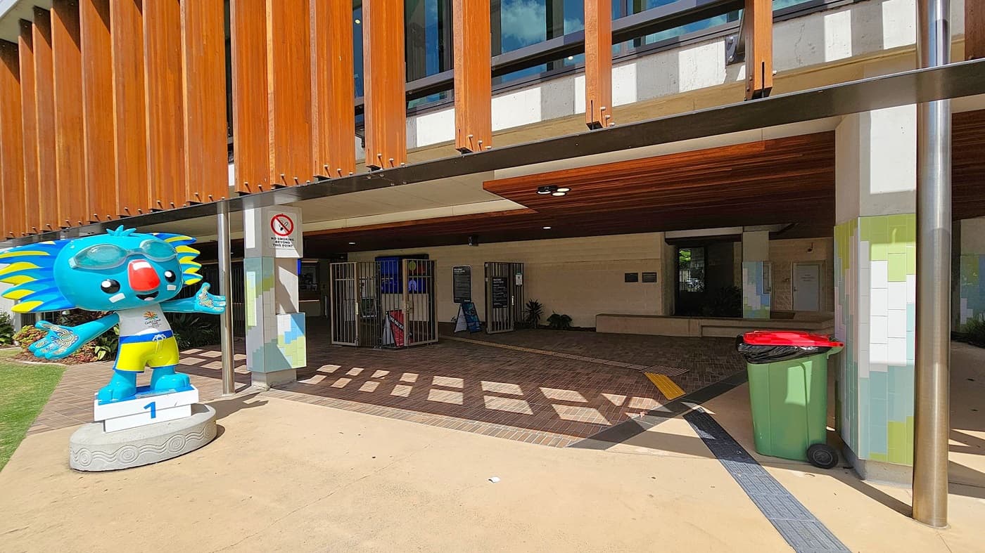 A colourful koala statue stands on the left, wearing goggles and yellow shorts, positioned on a platform marked with the number one. To the right, a green rubbish bin with a red lid is placed near a column with a tiled design. The background features a building entrance with wooden slats above, a no smoking sign, and a shaded area with brick flooring leading to a gated section.