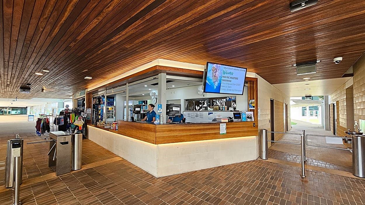 Reception area of an aquatic centre with a wooden ceiling and tiled floor. On the left, there are turnstiles and a display of swimwear and accessories. In the centre, a wooden reception desk with staff members and a digital screen displaying event information. The right side features a corridor leading to other areas, with a water fountain and more turnstiles. The area is well-lit with natural light coming from the background.