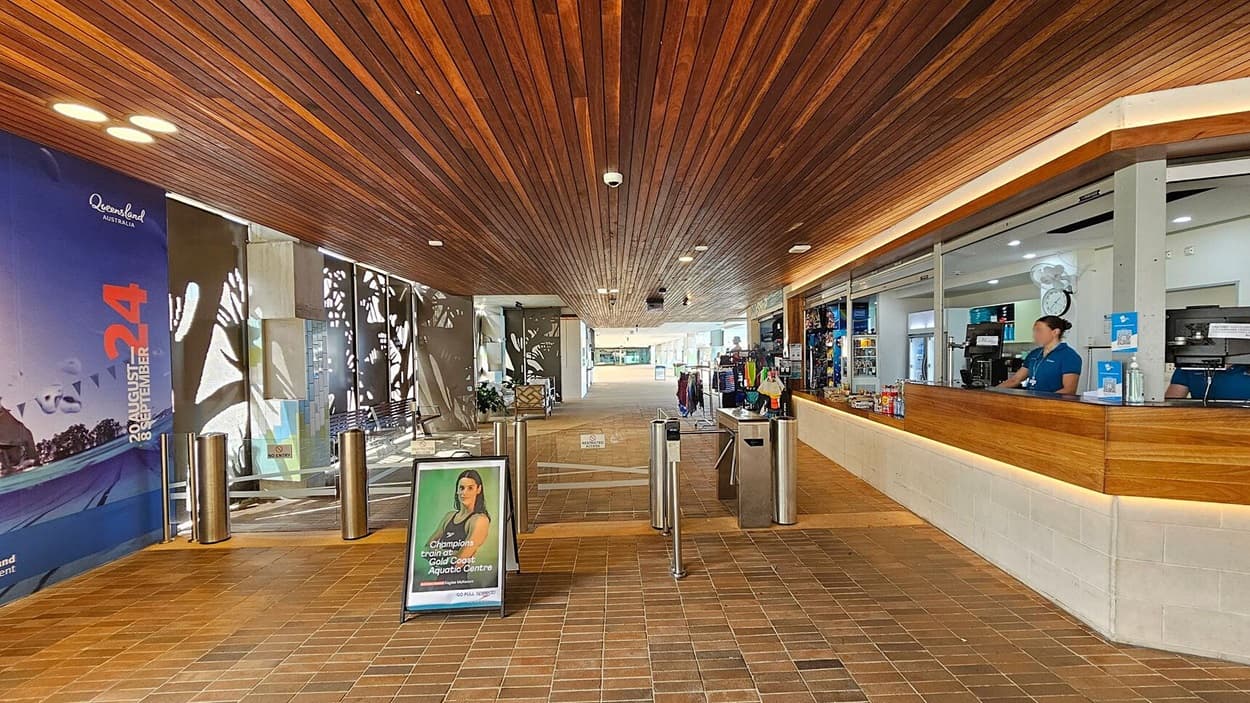 A spacious indoor area with a wooden ceiling and tiled floor. On the left, a large poster with "Queensland Australia" and event dates is displayed. In the centre, there are turnstiles and a promotional sign. On the right, a reception counter with a staff member behind it, surrounded by various items for sale. The background shows an open passage leading to another area.