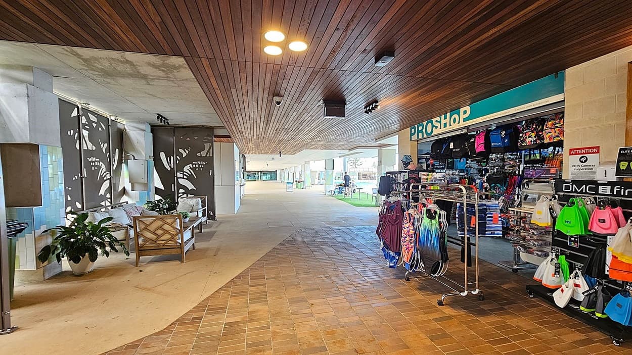 A corridor with a wooden ceiling and a tiled floor. On the left, there are decorative panels with plant motifs, a bench with cushions, and potted plants. On the right, a pro shop displays various colourful swimwear and accessories on racks and shelves. The shop has a sign indicating CCTV cameras are in operation. In the background, there is an open area with tables and chairs.