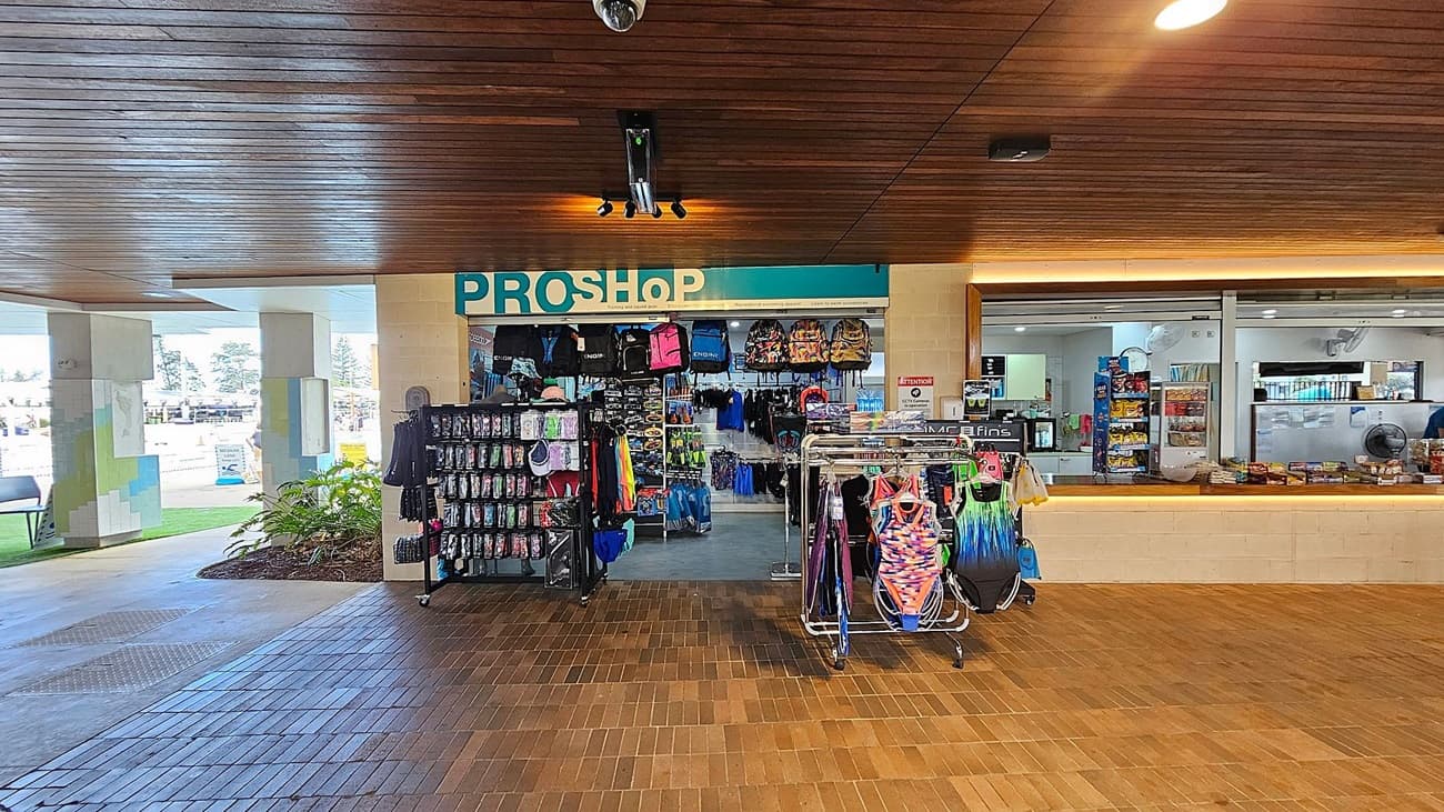 A pro shop with a wooden ceiling and tiled floor. In the centre, there are racks displaying swimwear, backpacks, and various accessories. To the right, a counter with snacks and drinks is visible, with a vending machine in the background. On the left, there are large windows and a small area with plants.