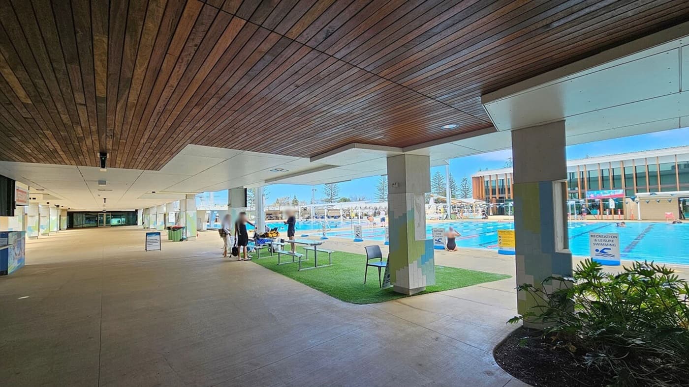 A covered walkway with a wooden ceiling and tiled floor leads to an outdoor swimming pool area on the right. The pool is surrounded by a grassy area with chairs and benches. People are walking and standing near the pool, which is divided into lanes. A building with large windows is visible in the background. Signs indicate areas for recreation and leisure swimming. Some plants are in the foreground near the walkway.