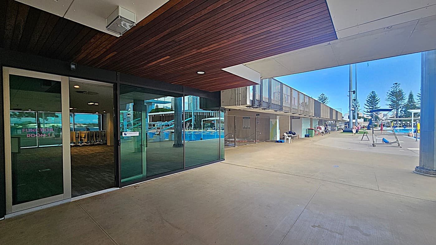 On the left, there is a glass door labelled "Function Room 1" leading to an interior space with chairs. The ceiling above is wooden. To the right, there is an outdoor area with a swimming pool, surrounded by a concrete floor. Several people are near the pool, and there are some chairs and equipment. In the background, tall trees and buildings are visible.