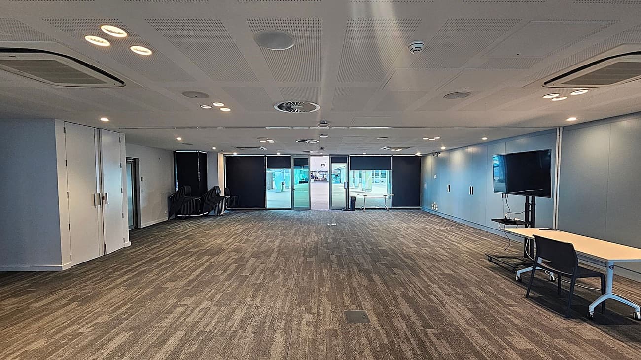 A spacious conference room with a patterned carpet floor. On the left, there are stacked chairs and a set of double doors. The back wall features large windows with black blinds, allowing natural light to enter. On the right, a flat-screen TV is mounted on a stand next to a table with a chair. The ceiling is equipped with recessed lighting and air conditioning vents.