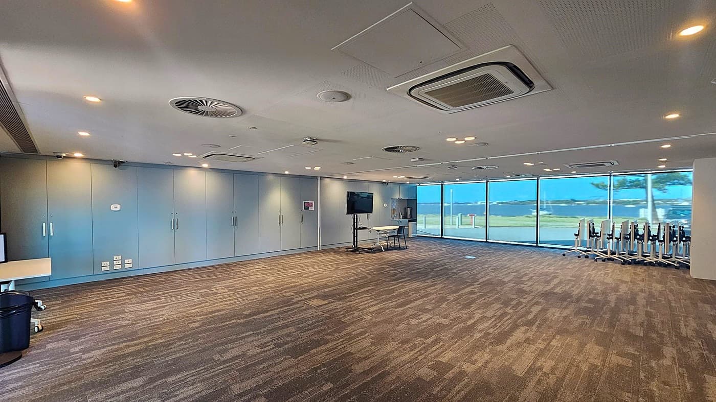 A spacious room with a patterned carpeted floor and a ceiling featuring multiple recessed lights and air conditioning vents. On the left, there are light blue storage cabinets with handles. In the centre, a large screen on a stand is positioned next to a small table and chair. On the right, a row of stacked chairs is visible. The back wall is made of large glass windows, offering a view of an outdoor area with a tree and open space.