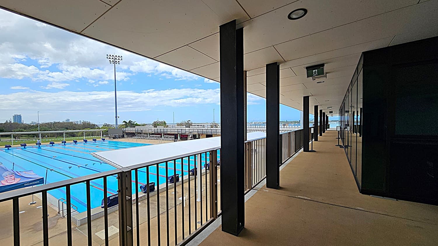 A covered walkway with a beige floor and ceiling, supported by black columns, runs along the right side. Glass panels line the right wall. On the left, a view of an outdoor swimming pool with blue lanes and starting blocks. Beyond the pool, there are bleachers and a tall floodlight. In the background, trees and distant buildings are visible.