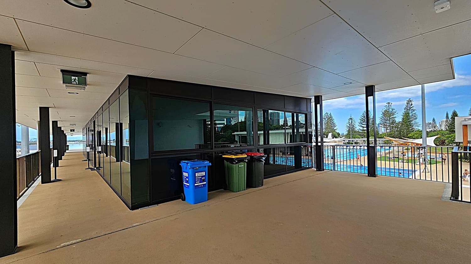 A corridor with a beige floor and a white ceiling featuring recessed lights and an exit sign. On the left, a glass wall reflects the opposite side, with a row of recycling bins in blue, yellow, green, and red. On the right, a railing overlooks an outdoor swimming pool area with people, surrounded by trees and buildings in the background.