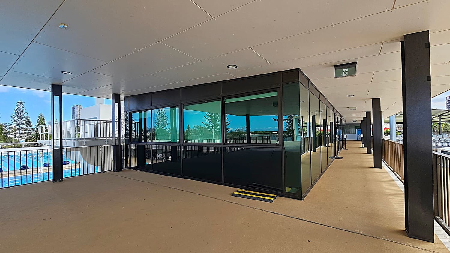A covered walkway with a beige floor and a white ceiling featuring recessed lights. On the right, there is a row of black-framed glass windows and doors. Black pillars support the structure, and a railing runs along the left side. Beyond the railing, a swimming pool with blue water is visible, along with some trees and a tall building in the background. An emergency exit sign is mounted on the ceiling.