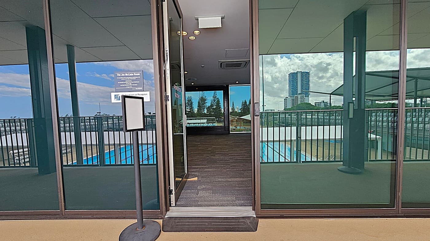 A glass door is open, leading into a room with a dark carpet. The ceiling inside has recessed lighting and an air vent. Outside, a balcony with a metal railing overlooks a swimming pool. Reflections on the glass show a tall building and cloudy sky. A sign is visible on the door, and a stand with a blank sign is positioned on the left. The floor outside is a light brown colour.