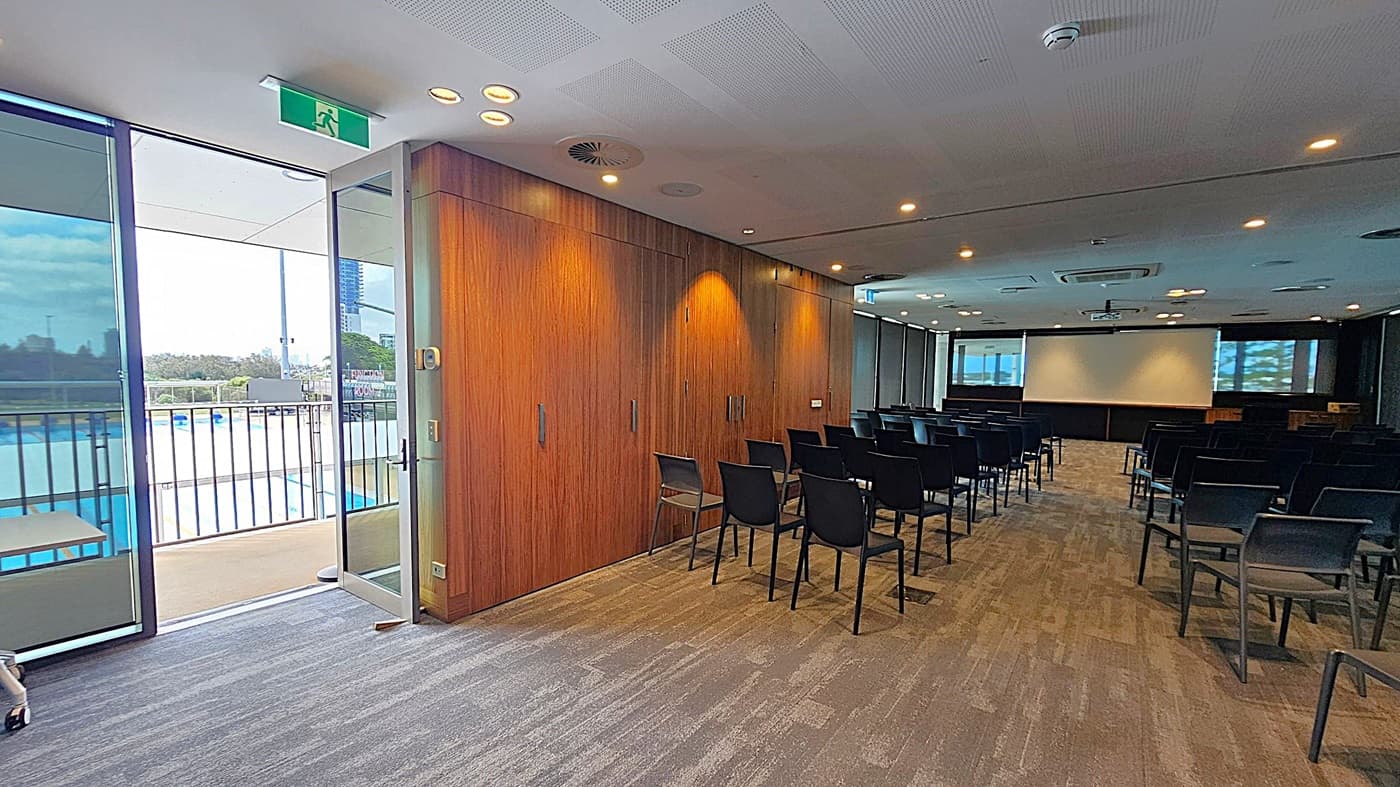 A conference room with a patterned carpet floor and a perforated ceiling with recessed lighting. On the left, there is a glass door leading to an outdoor area with a railing. The room has a wooden panelled wall with several black chairs arranged in rows facing a large projection screen at the far end. The room is well-lit with natural light coming through the windows on the left. An emergency exit sign is visible above the door.