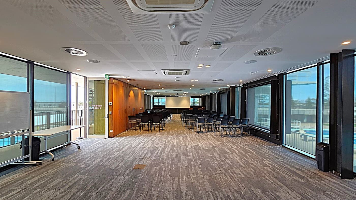 A conference room with a patterned carpet floor and a ceiling with recessed lighting and air vents. On the left, there's a whiteboard and a table on wheels near large windows with blinds. The right side also features large windows with blinds. In the centre, rows of black chairs face a projection screen at the far end. The walls are a mix of wood panelling and glass. A door labelled "Function Room 2" is on the left wall.