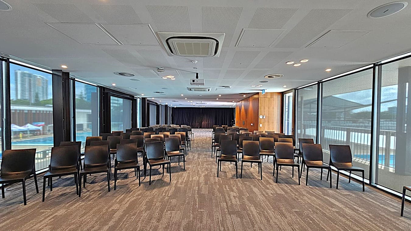 A conference room with rows of black chairs facing a stage area at the back. The room has large floor-to-ceiling windows on both sides, allowing natural light to enter. The ceiling is equipped with air conditioning vents and a projector. The floor is carpeted in a multicoloured pattern. The walls on the right are wooden, and the left side shows an outdoor view with buildings and a pool.