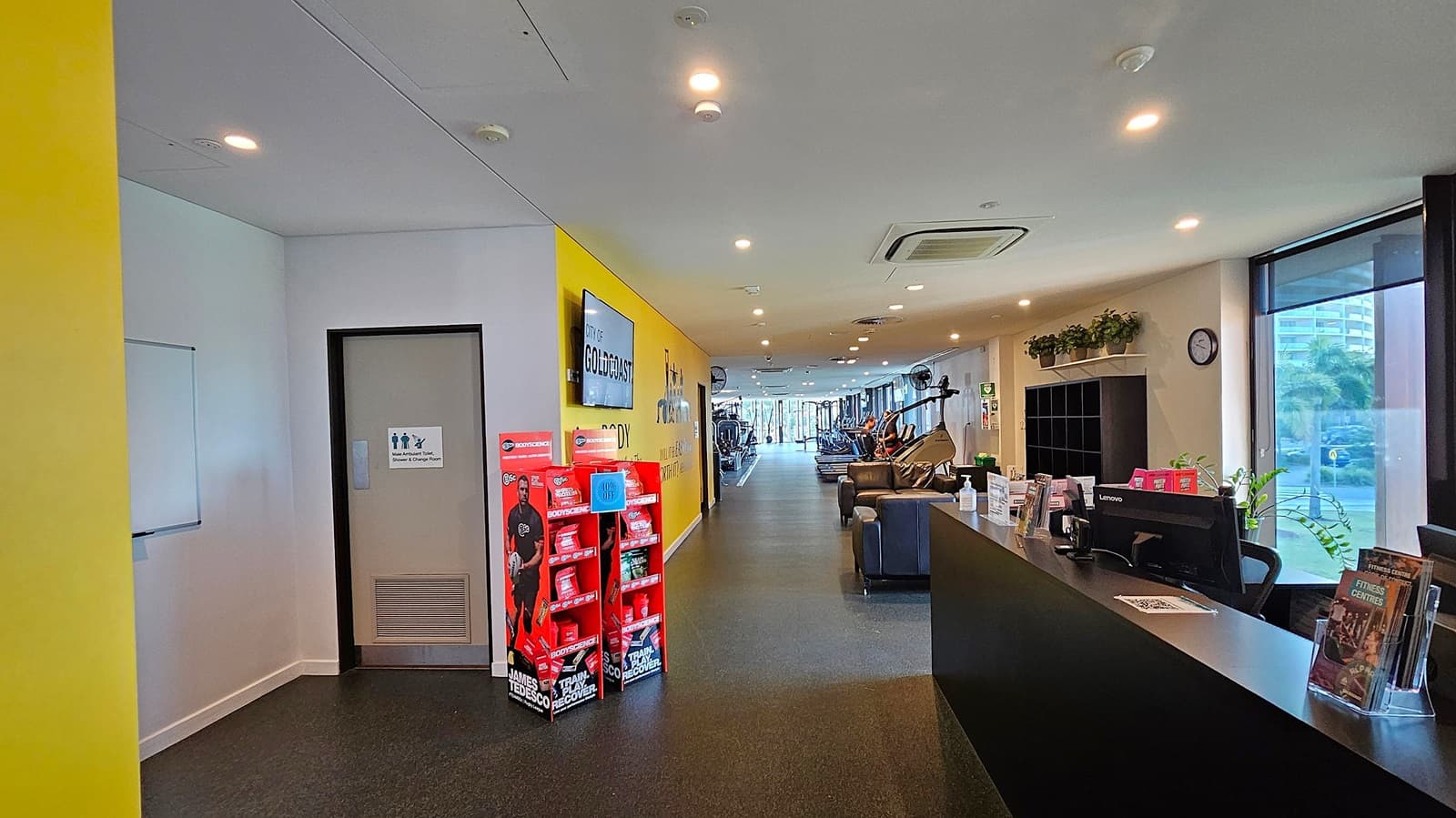 A covered outdoor area with a ceiling featuring recessed lights and a security camera. On the left, there's a door marked "Fire Hydrant" and "Fire Hose Reel." In the centre, a glass door leads to an indoor space with visible people. A lift is adjacent to the door. On the right, a railing encloses a ramp, and several bins are placed nearby. The background shows a spacious open area with a canopy and trees in the distance.