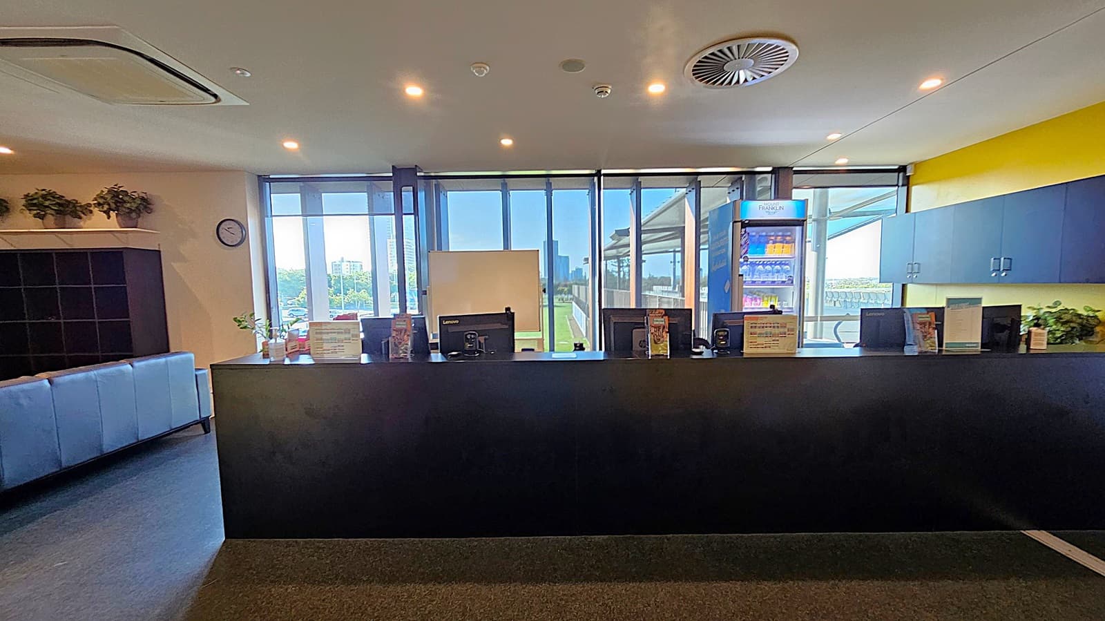 A reception area with a black counter in the foreground, featuring computer monitors and informational brochures. Behind the counter, large windows provide a view of a green outdoor area. To the right, a refrigerated display with drinks is visible. On the left, a wall clock and potted plants are placed above a shelving unit. The ceiling has recessed lighting and air conditioning vents. The floor is dark and smooth.