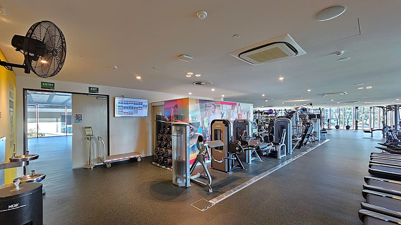 A spacious gym interior with a variety of exercise equipment. On the left, there's a wall-mounted fan, a water fountain, and an entry/exit door with a digital screen above it. In the centre, there are weight racks and various gym machines. The right side features a row of treadmills. The floor is covered with dark rubber matting, and the ceiling has multiple recessed lights and air conditioning vents. Large windows in the background allow natural light to enter.