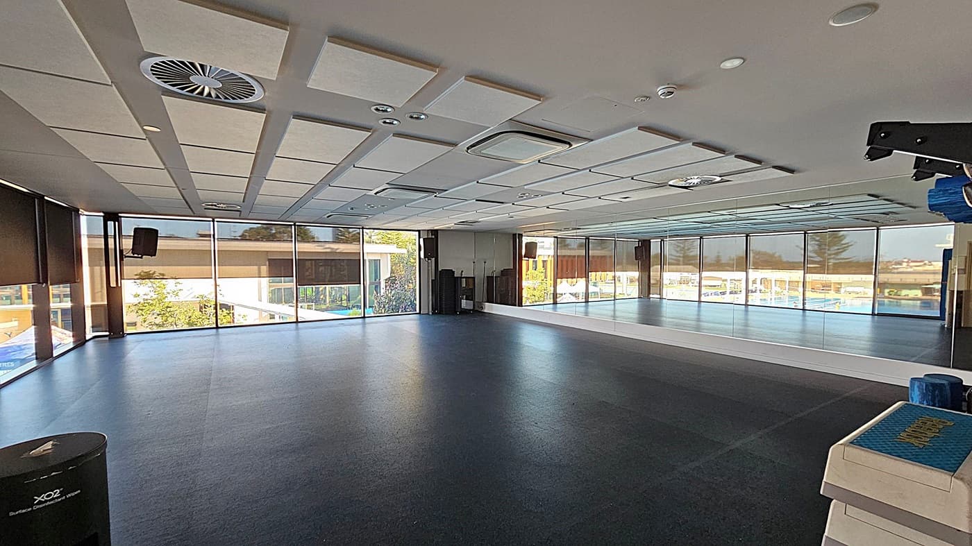 A spacious room with large floor-to-ceiling windows on the left and back walls, allowing natural light to fill the space. The right wall is covered with mirrors, reflecting the room's interior. The ceiling features a grid pattern with recessed lighting and air vents. The floor is dark and smooth, suitable for exercise or dance. In the foreground on the right, there are stacked exercise mats. A speaker is mounted on the left wall.