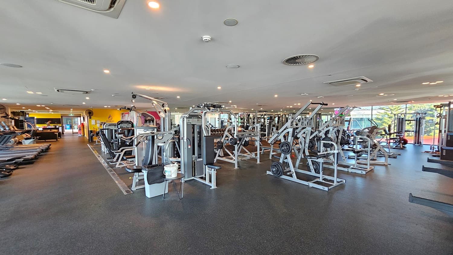 A spacious gym with various exercise machines. On the left, there are several treadmills lined up. In the centre, there are weight machines and benches. To the right, more weightlifting equipment is visible. The floor is dark and the ceiling is white with recessed lighting and air conditioning vents. Large windows in the background allow natural light to enter, with a view of trees outside.