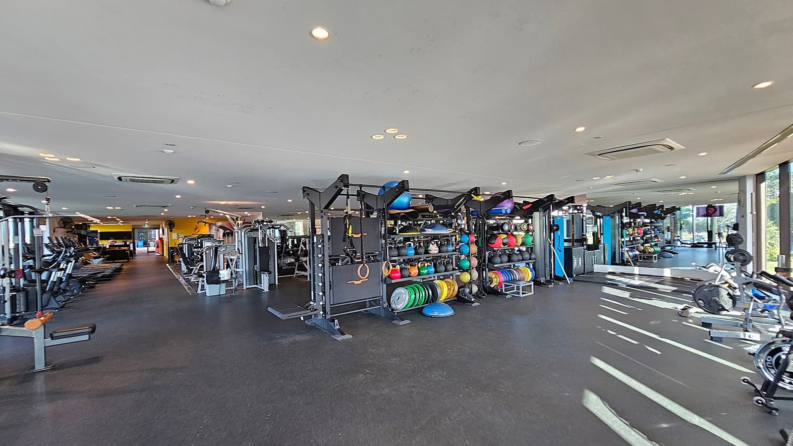 A spacious gym with a variety of equipment. On the left, there are treadmills and weight machines. In the centre, racks hold colourful medicine balls, kettlebells, and weight plates. On the right, there are exercise bikes and a mirrored wall reflecting the equipment. The floor is dark and the ceiling is light with recessed lighting. Large windows on the right let in natural light.