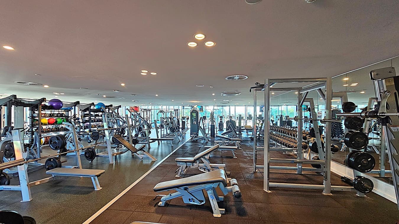 A spacious gym with various exercise equipment. On the left, there are racks of colourful medicine balls and weight plates. In the centre, several benches and weight machines are arranged neatly. On the right, a squat rack with weight plates is visible. The floor is covered with rubber mats, and the ceiling has recessed lighting. Large windows in the background allow natural light to fill the room.