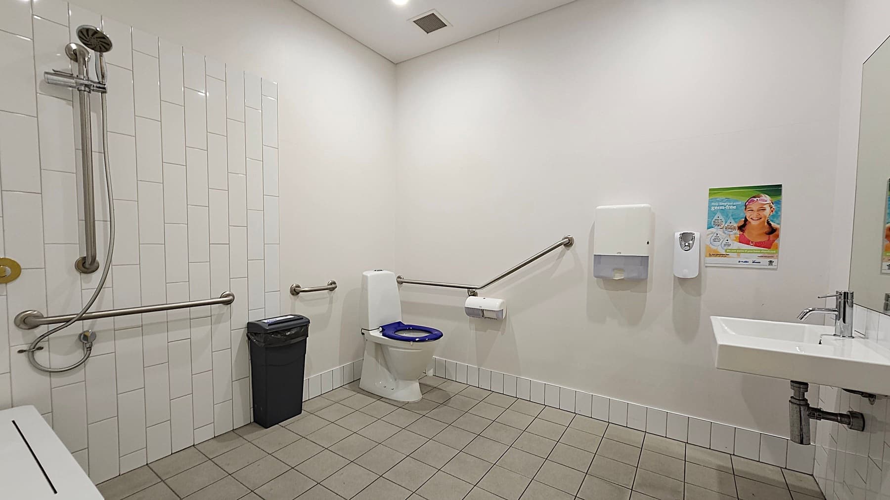 A public bathroom with a green-tiled floor and white walls. On the left, there is a shower with a handheld showerhead and metal grab bars. In the centre, a toilet with a blue seat is equipped with grab bars on both sides. A black bin is next to the toilet. On the right, a white sink with a metal tap is mounted on the wall. Above the sink, a paper towel dispenser and a soap dispenser are installed. A poster is visible on the wall near the sink.