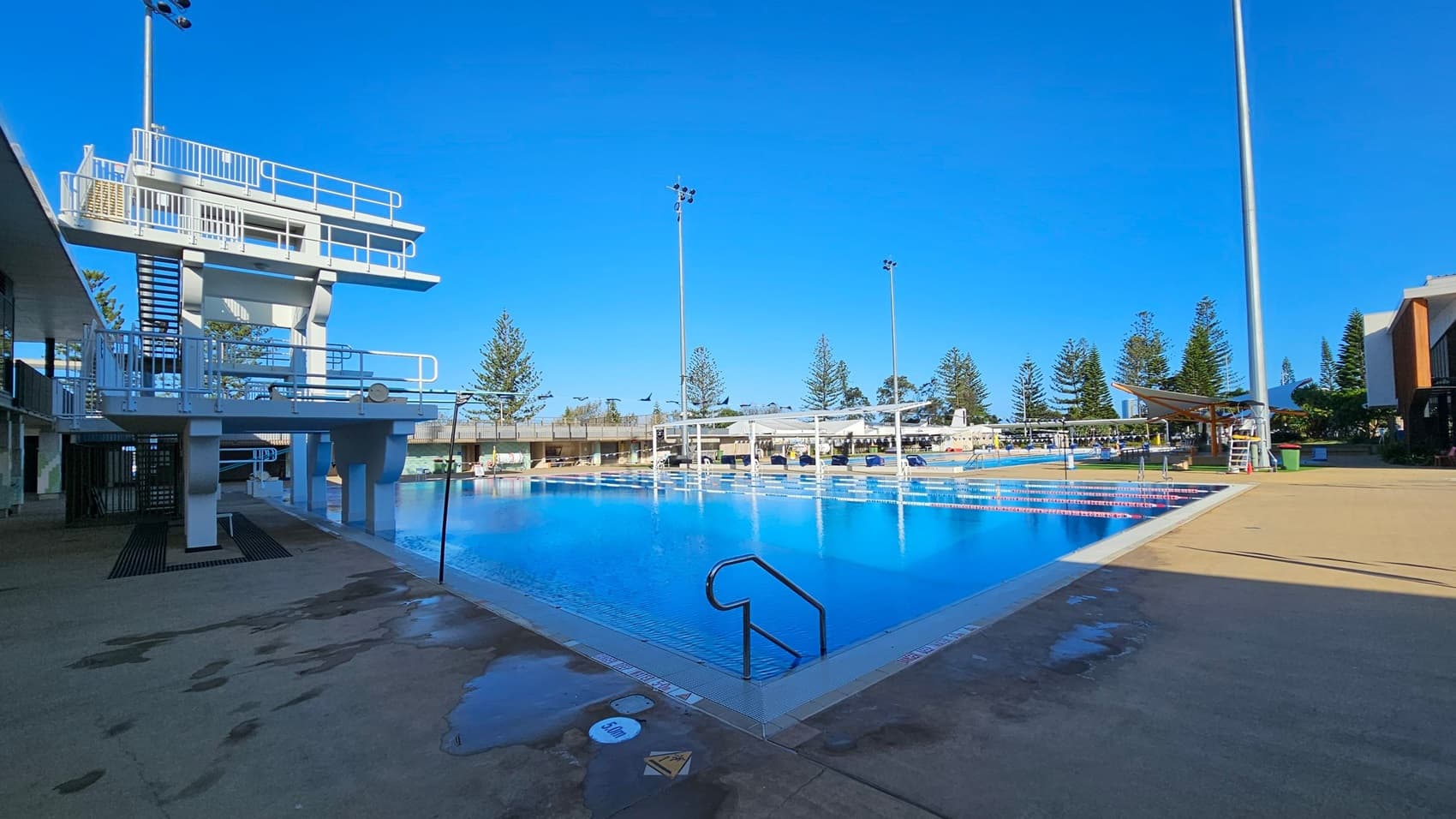 A large outdoor swimming pool with clear blue water is in the centre. On the left, there is a tall diving platform with multiple levels and stairs. The pool is surrounded by a concrete deck with some wet patches. In the background, there are tall trees and several light poles. On the right, there is a shaded seating area and a lifeguard chair. The sky is clear and blue.