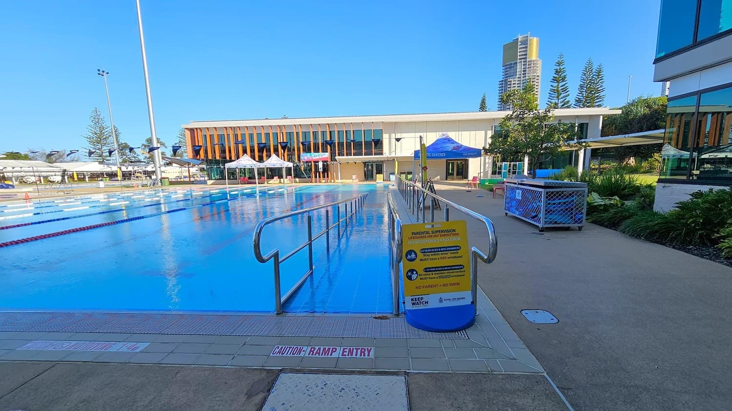 An outdoor swimming pool with clear blue water is in the centre, surrounded by a concrete deck. On the right, a row of white chairs is under a shaded canopy. Tall buildings and pine trees are visible in the background. The pool has lane dividers and a "Caution Deep Water" sign. A metal ladder is on the left side of the pool.