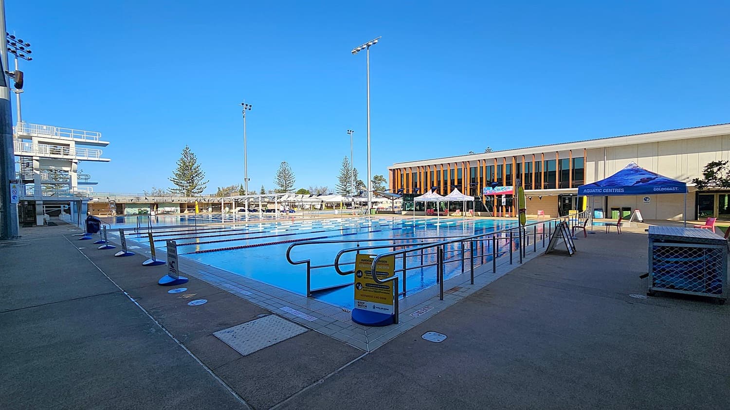 An outdoor swimming pool with lane dividers is on the left, with a ramp entry and a caution sign. On the right, there's a paved area with a sign about parental supervision. In the background, a modern building with large windows and vertical wooden slats is visible, along with several tents and a tall building. Trees and greenery are also present around the area.