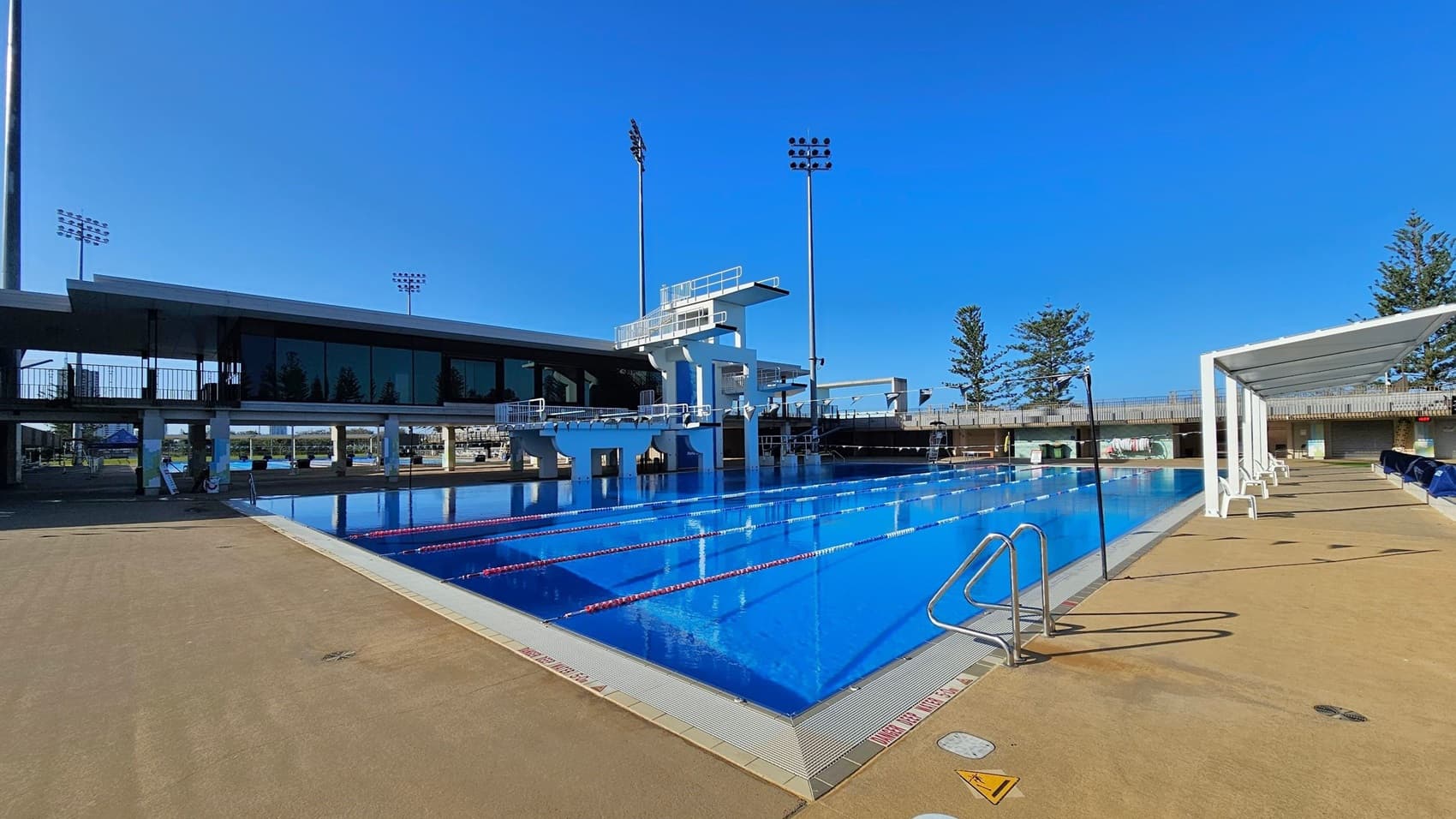 An outdoor swimming pool with clear blue water and lane dividers. On the left, there's a diving platform with multiple levels. The pool is surrounded by a beige deck area. On the right, a row of white chairs is shaded by a canopy. In the background, a modern building with large windows and tall floodlights is visible. Trees are scattered around the perimeter, and the sky is clear.