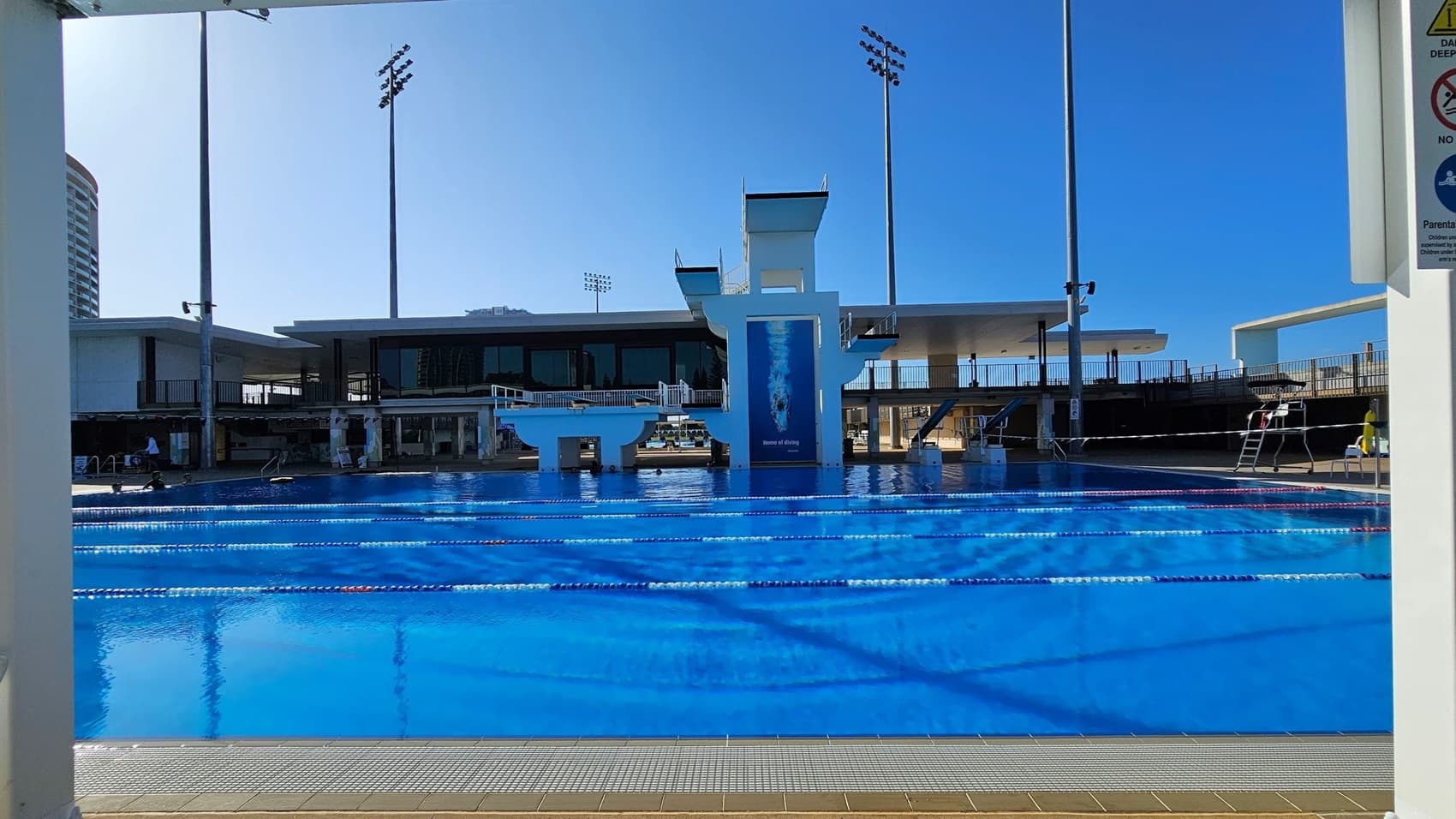 An outdoor swimming pool with clear blue water and lane dividers. In the background, there is a diving platform with multiple levels and a large poster of a diver. The pool area is surrounded by a modern building with large windows and a flat roof. Tall floodlights are visible, and there are signs on the right side. The pool deck is tiled, and a lifeguard chair is positioned near the pool.