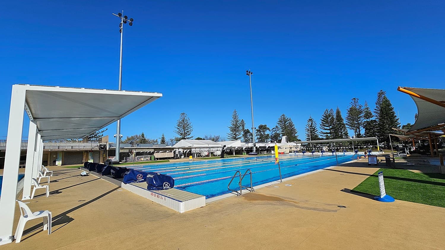 An outdoor swimming pool with clear blue water and lane dividers stretches across the centre. On the left, there are white plastic chairs under a shaded structure. On the right, a grassy area with more shade structures. Tall trees line the background, and a tall light pole stands near the pool. The ground is paved with a light brown surface.