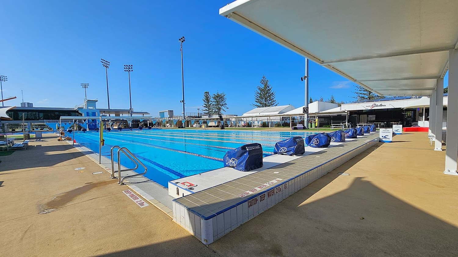 An outdoor swimming pool with clear blue water and lane dividers stretches across the centre. On the right, a shaded walkway with signs and covered equipment. The left side has a concrete deck with a metal pool ladder. In the background, tall floodlights and trees are visible, along with a building labelled "Cafe." The ground is a mix of concrete and tiled surfaces.