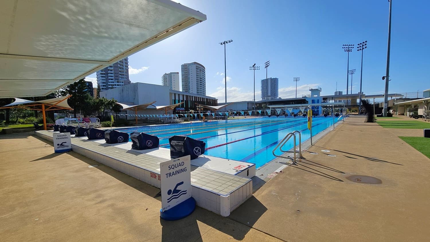 An outdoor swimming pool with clear blue water is in the centre, surrounded by a concrete deck. On the left, there are shaded areas with white canopies and a sign reading "Squad Training." Several covered starting blocks line the pool's edge. In the background, tall buildings and floodlights are visible. The right side features a grassy area and more concrete decking. The sky is clear and sunny.
