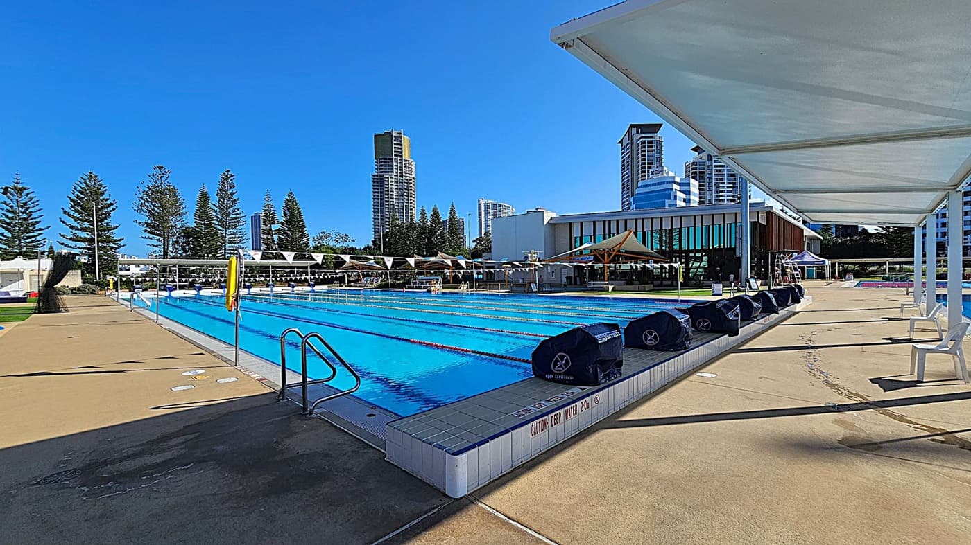 An outdoor swimming pool with clear blue water is in the centre, surrounded by a concrete deck. On the right, a row of white chairs is under a shaded canopy. Tall buildings and pine trees are visible in the background. The pool has lane dividers and a "Caution Deep Water" sign. A metal ladder is on the left side of the pool.