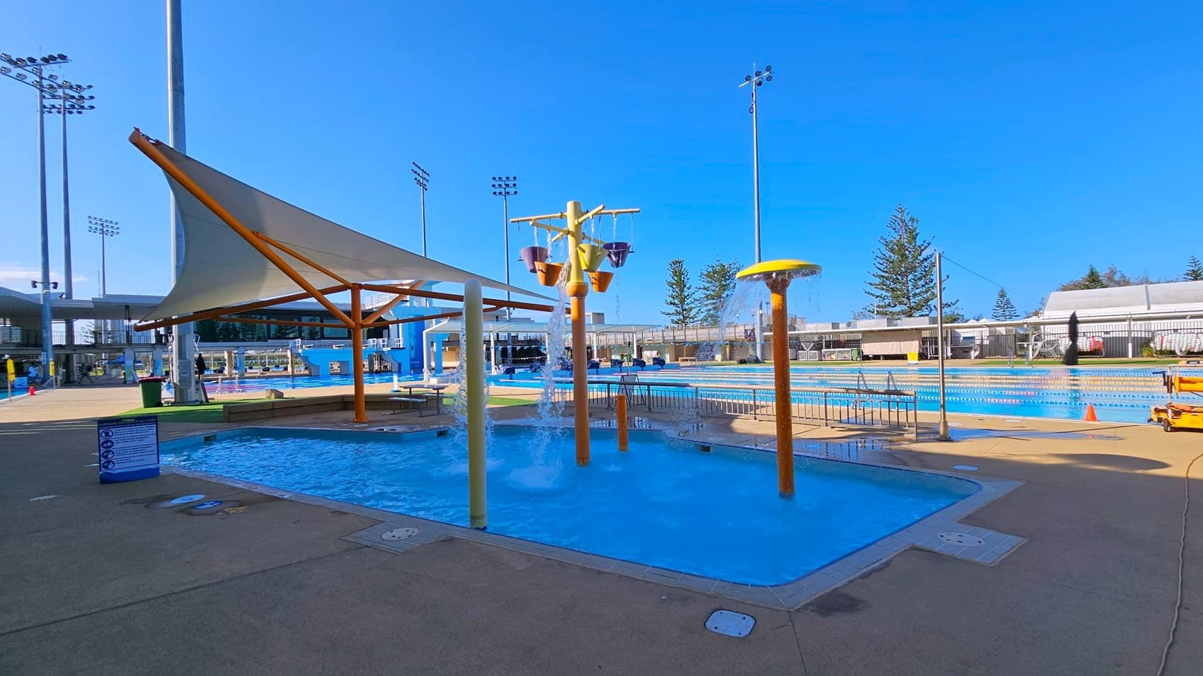 A swimming pool area with a shallow play pool in the foreground, featuring water fountains and colourful buckets. To the left, a large shade structure with a white canopy supported by wooden beams. In the background, a larger lap pool with lane dividers and a few trees. Tall floodlights are visible around the area, and the ground is paved with concrete.