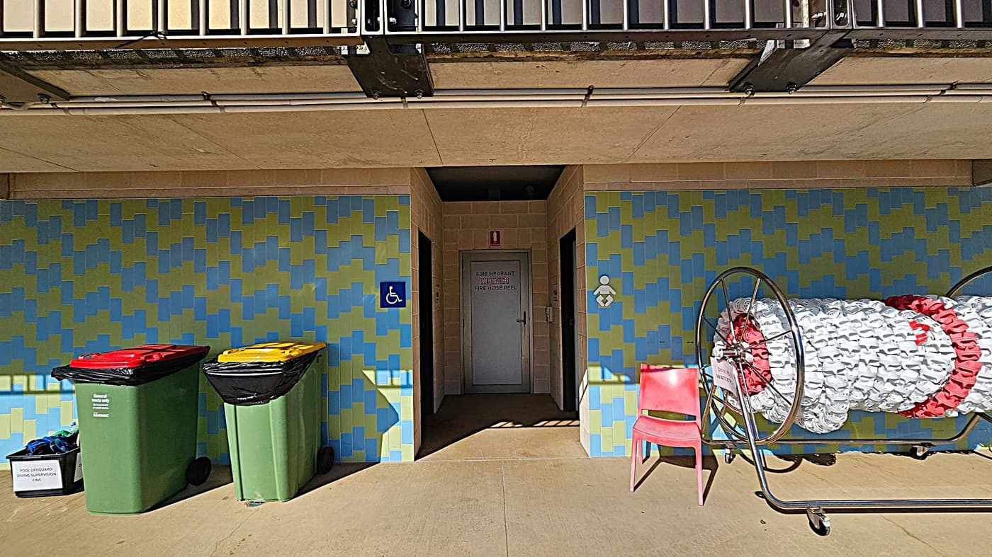 A building exterior with a blue and green patterned wall. In the centre, a door marked "Fire Hydrant" is flanked by two signs indicating accessible facilities. On the left, two green bins with red and yellow lids stand next to a box labelled "Pool Lifeguard Diving Session Fins." On the right, a red chair is beside a large reel of white and red material. The ceiling is beige with visible pipes, and a metal railing is above.