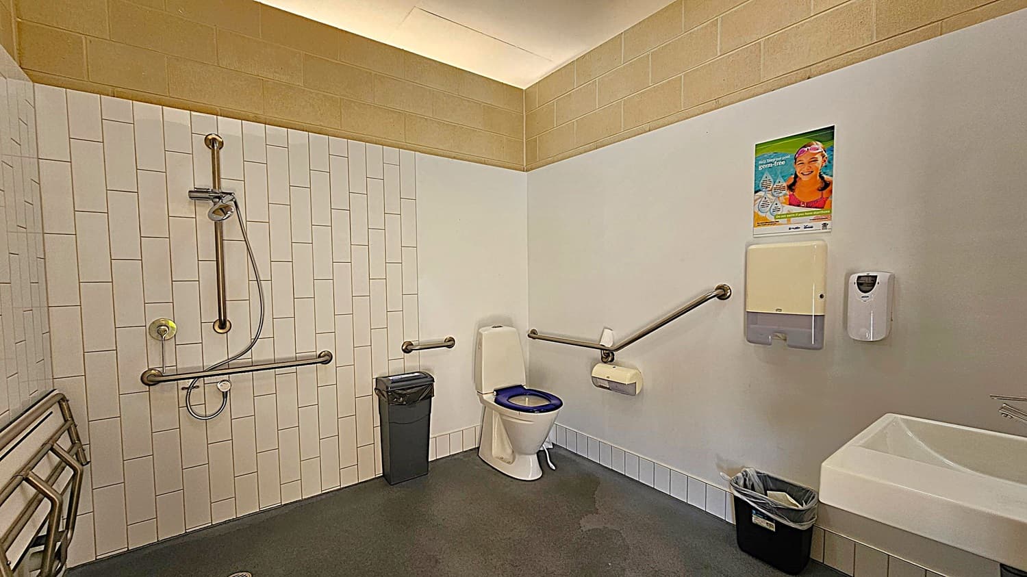 Accessible bathroom with a shower on the left, featuring a handheld showerhead and grab bars. A toilet with a blue seat is in the centre, also equipped with grab bars. A bin is next to the toilet. On the right, there's a sink, a paper towel dispenser, and a soap dispenser. A poster is above the dispenser. The floor is dark, and the walls are tiled with a mix of white and beige tiles.