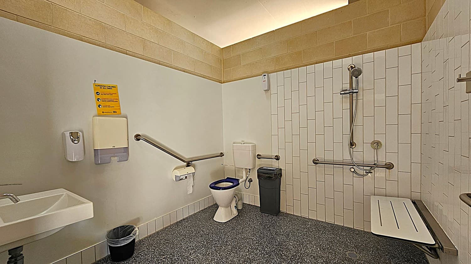 A public accessible bathroom with a white sink on the left, accompanied by a soap dispenser and paper towel holder. A small bin is on the floor. In the centre, a toilet with a blue seat is flanked by grab bars, with a waste bin nearby. On the right, a shower area features a handheld showerhead, grab bars, and a foldable seat. The walls are tiled, and the floor has a speckled pattern. The ceiling is light-coloured with a recessed lighting feature.