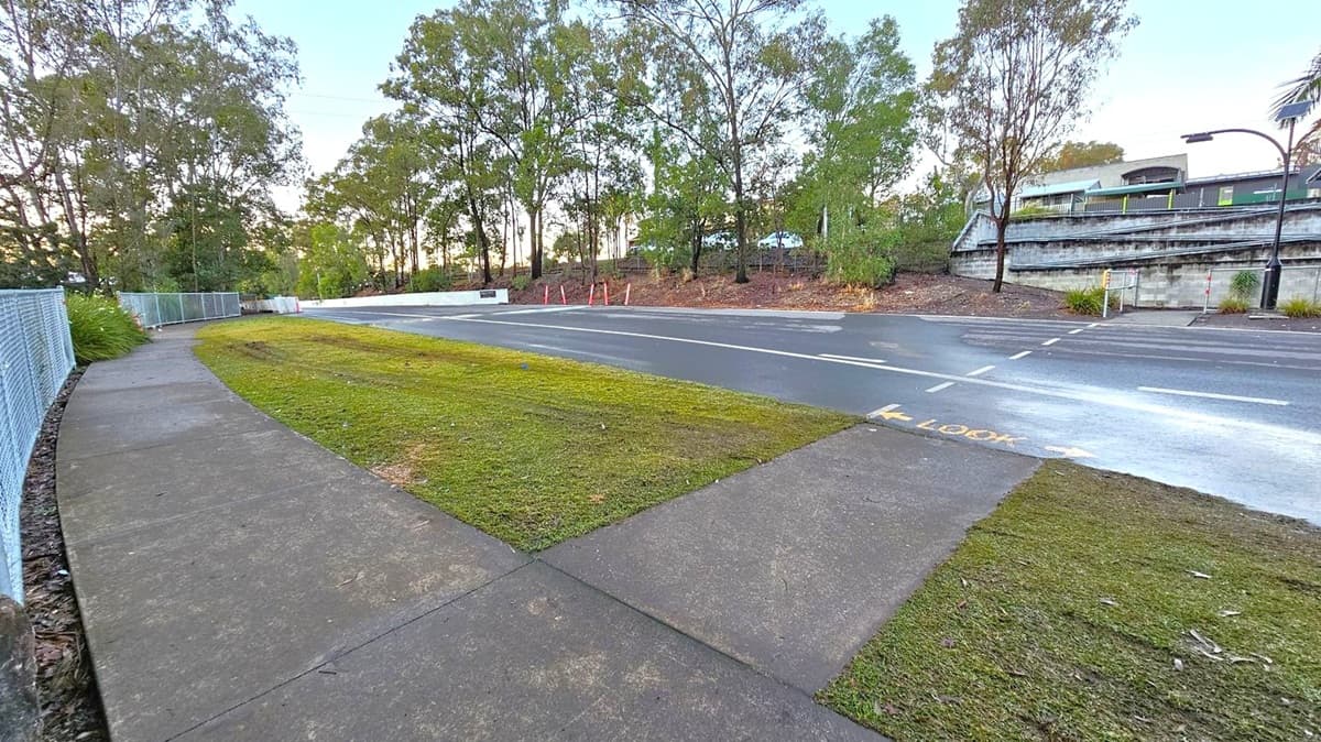 A curved concrete footpath runs along the left side, bordered by a chain-link fence and greenery. In the centre, a grassy area separates the path from a road. The road has a pedestrian crossing marked with "LOOK" in yellow. On the right, a train is visible on elevated tracks, supported by a concrete wall. Tall trees line the background, providing a natural backdrop.