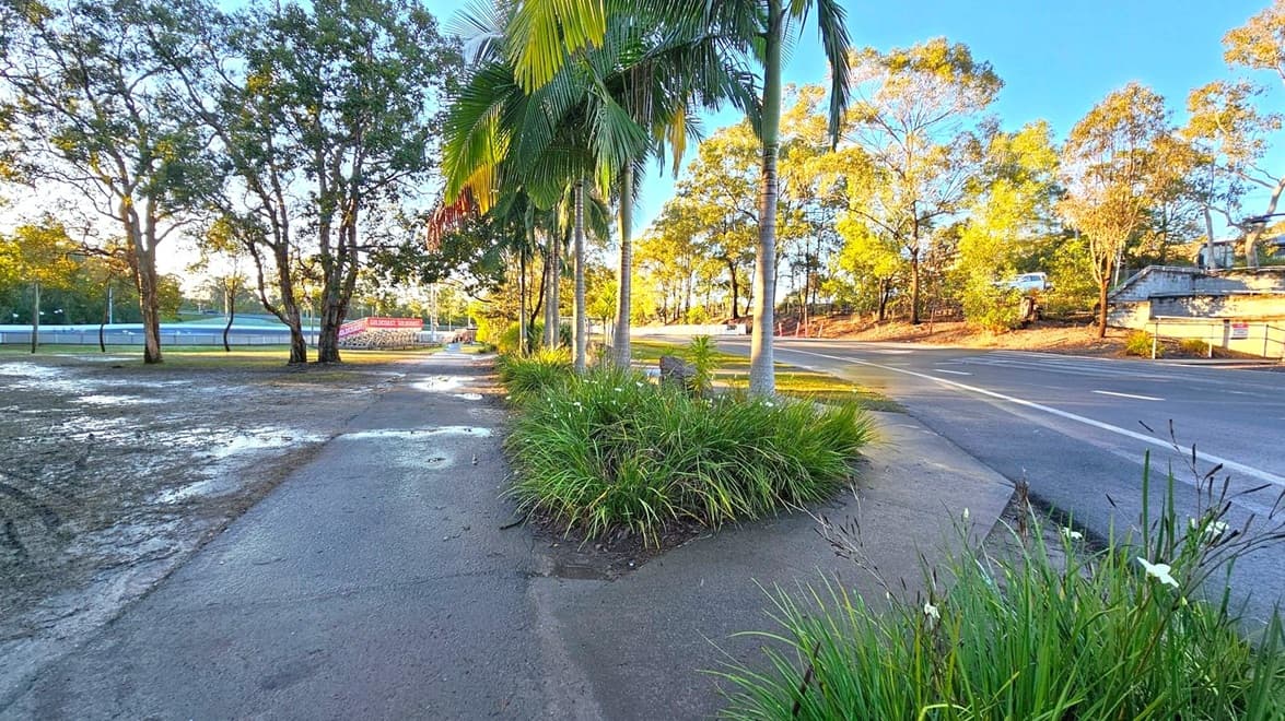 A paved path runs through the centre, bordered by lush green plants and tall palm trees. On the left, there are large trees and a grassy area with puddles, leading to a fenced sports field with a sign. On the right, a road curves gently, lined with more trees and a parked vehicle. The scene is bathed in warm sunlight, casting long shadows.