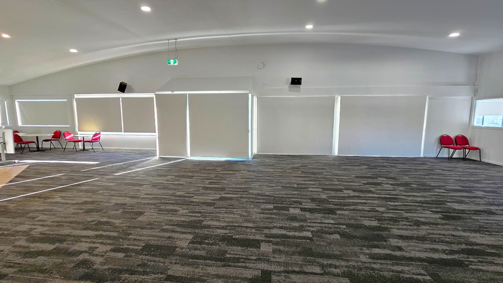 A spacious room with a patterned carpet floor and a white ceiling with recessed lighting. On the left, there are several red chairs around small tables near large windows with blinds. In the centre, the wall has large, closed blinds. On the right, two red chairs are placed against the wall near another window. The room has a modern and minimalistic design.