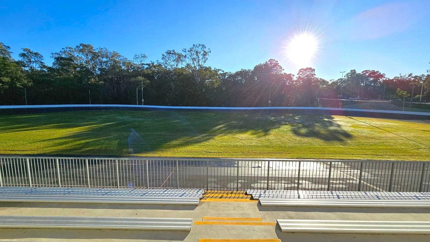 Bleachers in the foreground with metal railings and steps leading down to a grassy field. The field is surrounded by a white barrier, and beyond it, there are tall trees casting long shadows. The sun is low in the sky, creating a bright glare and illuminating the scene. The sky is clear and blue.