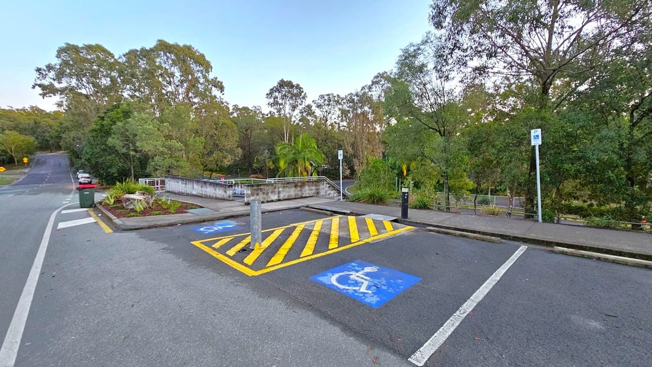 A parking area with two marked disabled parking spaces in the foreground, featuring blue wheelchair symbols and yellow diagonal lines. A small garden with plants and a green bin is on the left. A road extends into the distance on the left side. On the right, a footpath runs alongside a railing, with dense trees and greenery in the background. A parking sign is visible near the trees.